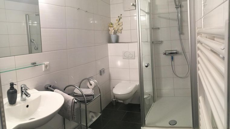 A modern bathroom with a shower, a sink, and a toilet. The walls are tiled in white, and there is a plant on the shelf.