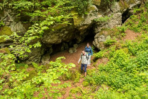 Twee wandelaars komen uit een grottingang van een massieve rotswand naar buiten in een dicht loofbos.