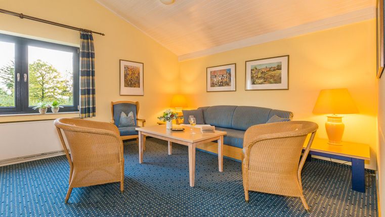 Ein gemütliches Wohnzimmer mit einem blauen Sofa und zwei Korbsesseln. Der Raum hat gelbe Wände und ist gut beleuchtet, mit Bildern an der Wand.