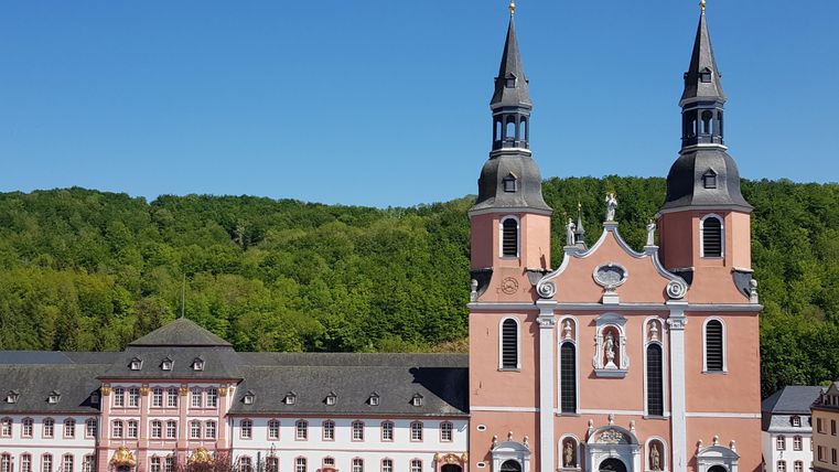 Eine beeindruckende Kirche mit zwei Türmen und einer roten Fassade. Im Hintergrund sind grüne Hügel und eine ruhige Umgebung sichtbar.