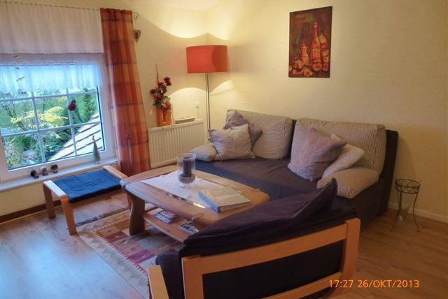 Ein gemütliches Wohnzimmer mit einer großen Couch, einem Couchtisch und einem Fenster mit Blick ins Grüne. Die Wände sind hell, und eine Stehlampe sorgt für angenehmes Licht.