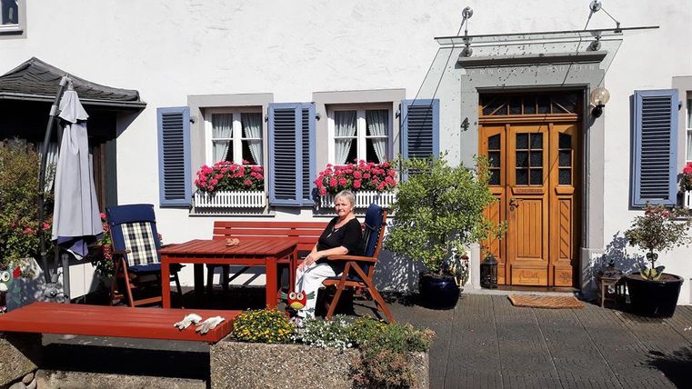 A woman sits relaxed on a terrace in front of a beautiful house with blue shutters and blooming plants. The wooden table and chairs invite you to linger.