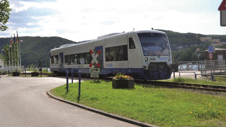 A modern train passes by a stop. The surroundings are green and rural, with gentle hills in the background.