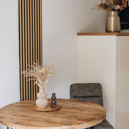 Ein runder Holztisch mit modernen Stühlen und einer Vase mit Trockenblumen. Die Wände sind schlicht und hell, was einen einladenden Raum schafft.