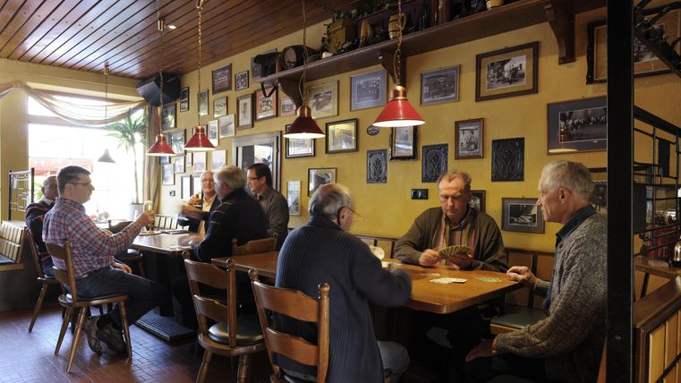 Eine gemütliche Kneipe mit mehreren Männern, die am Tisch sitzen und Karten spielen. Die Wände sind mit vielen Fotos dekoriert.
