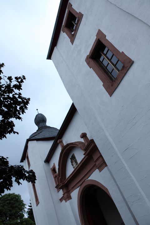 Wit gebouw met rode raamkozijnen en een toren met een gebogen dak. De lucht is bewolkt, een boom is op de voorgrond zichtbaar.
