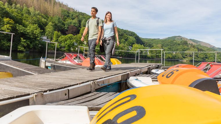 Ein Paar spaziert auf einem Steg bei einem Bootsverleih am Wasser. Im Hintergrund sind grüne Hügel und farbige Boote zu sehen.