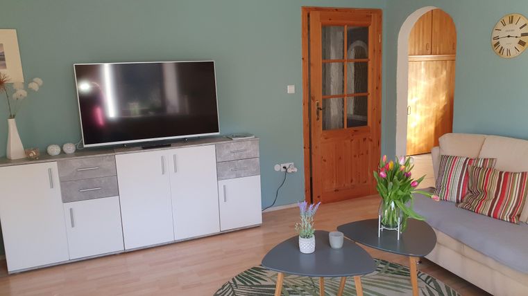 Een gezellige woonkamer met een televisie, een lichte bank en een tafel. De ruimte heeft groene muren en planten op de tafel, die een uitnodigende sfeer creëren.