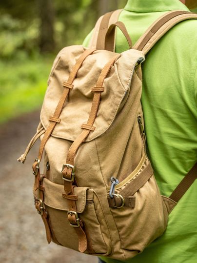 A beige backpack with brown leather straps, worn by a person in a green T-shirt. The scene shows a forest path in the background.