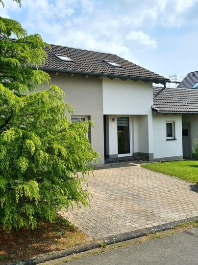 Ein modernes Haus mit einem gepflasterten Gehweg und Rasenflächen. Einige Bäume bieten Schatten vor dem Eingang.