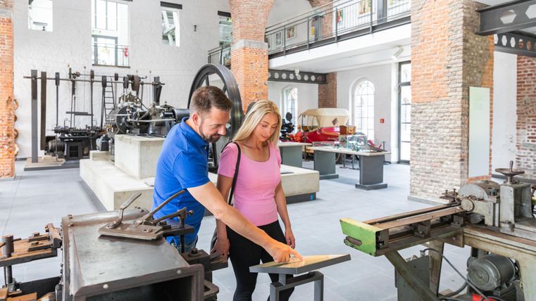 Two people are jointly examining a document in a museum for old machines. The room is well-lit and features historical devices.