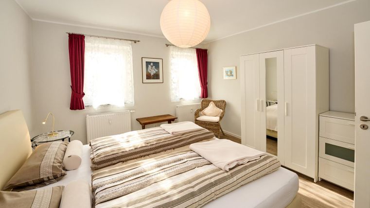 Een lichte slaapkamer met een groot bed en gestreepte beddengoed. Er zijn ramen met witte gordijnen en een gezellige stoel in de hoek.