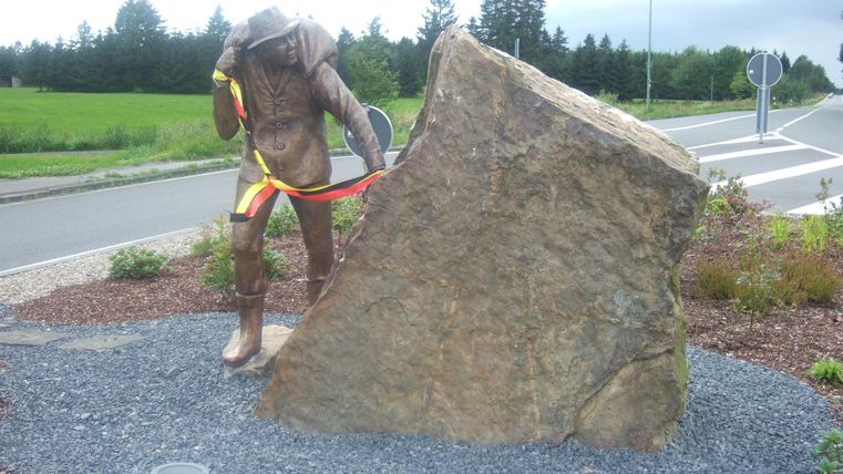 Eine Statue eines Arbeiters, der an einem großen Stein arbeitet. Die Umgebung ist grün mit einigen Sträuchern und einer Straße im Hintergrund.