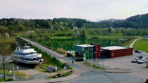 Ein ruhiger Hafen mit einem Schiff und einem modernen Gebäude in der Nähe eines klaren Gewässers. Umgeben von grünen Wäldern und Wiesen.