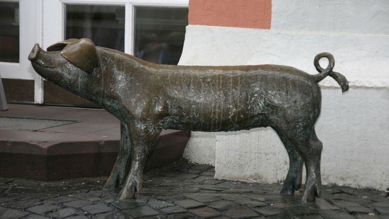 Eine bronzene Statue eines Schweins steht auf dem Boden. Sie hat eine eindeutige, detaillierte Form und ist in einer urbanen Umgebung positioniert.