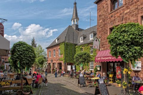 Eine malerische Gasse mit historischen Gebäuden und einem kleinen Café. Menschen sitzen draußen und genießen die Atmosphäre unter den grünen Bäumen.