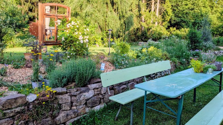 A beautiful garden with a stone wall and colorful flowers. A table and benches invite you to relax.