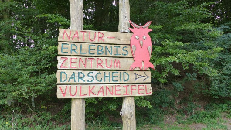 Een houten bord met meerdere richtingpijlen en kleurrijke letters. Het toont de weg naar het natuurbelevingscentrum Darscheid in de Vulkaneifel.