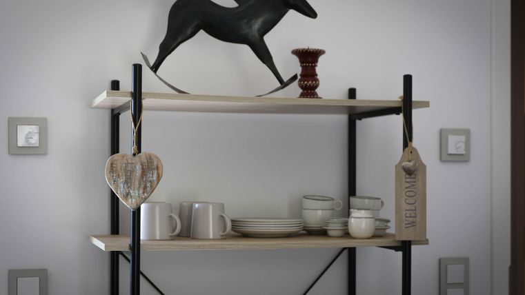A modern shelf with a black horse decoration piece on top. Below are cups, plates, and a heart-shaped ornament as well as a welcome sign.