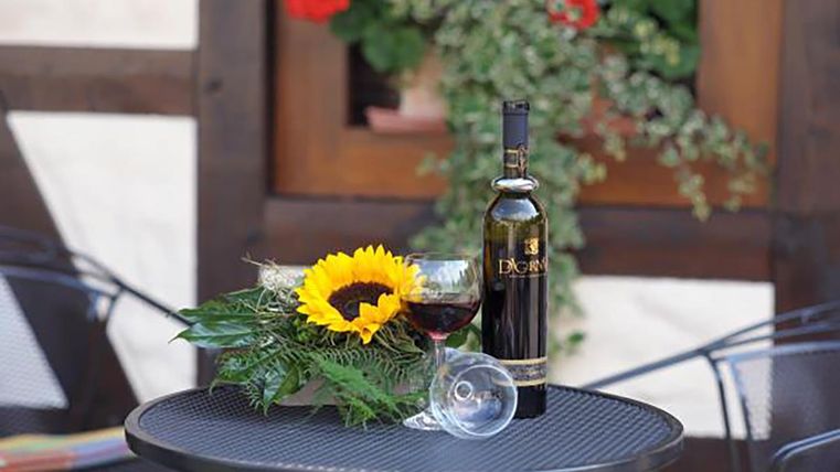 Ein Tisch im Freien mit einer Flasche Wein, einem Glas Wein und einer Sonnenblume. Im Hintergrund sind blühende Pflanzen zu sehen.