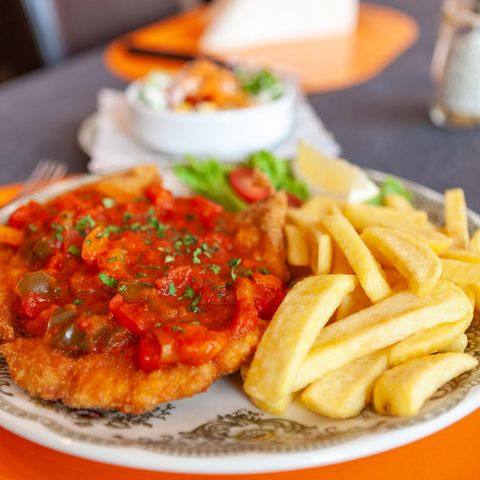 Ein Teller mit Schnitzel, bedeckt mit Paprikasauce, serviert mit Pommes frites und einem Salat im Hintergrund.