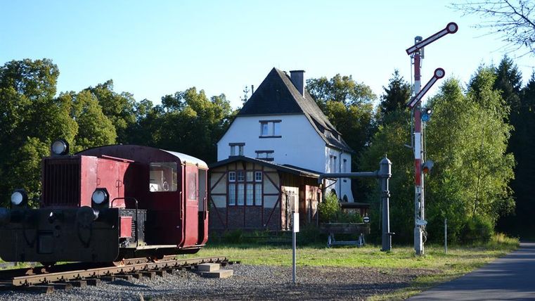 Eine alte Lokomotive steht auf Gleisen vor einem weißen Haus. Im Hintergrund sind Bäume und ein Stellwerkssignal zu sehen.