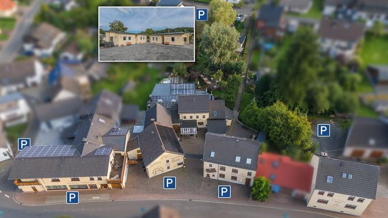 Eine Luftaufnahme von mehreren Gebäuden mit Parkplätzen. Im oberen Bereich ist ein Bildausschnitt eines größeren Bauprojekts zu sehen.