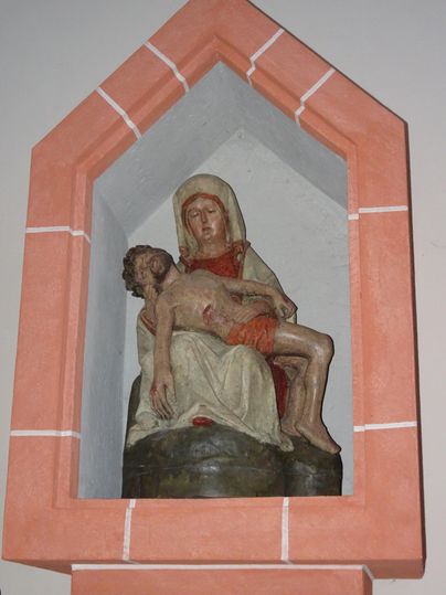 Een standbeeld van de Pietà toont Maria die het levenloze lichaam van Jezus vasthoudt. De sculptuur is in een nis geplaatst en heeft een eenvoudige, neutrale achtergrond.