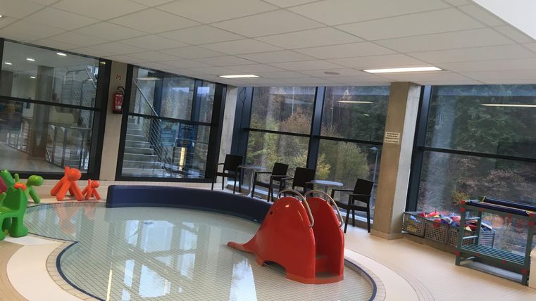 Een lichte kamer met een ondiep bassin en grote vensters. Er zijn kleurrijke speeltoestellen in de buurt van het water.