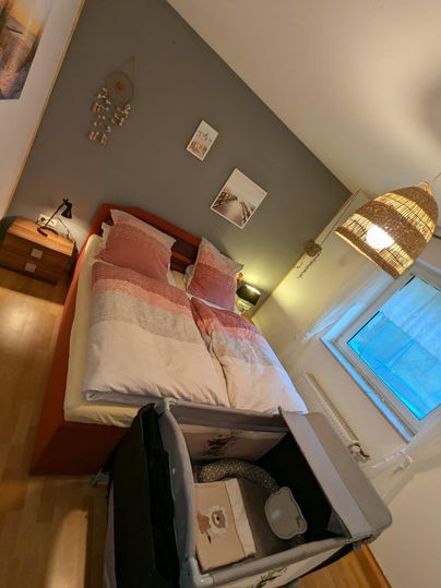 A modern bedroom with a double bed and decorative pillows. The room also features a crib and a window with natural light.