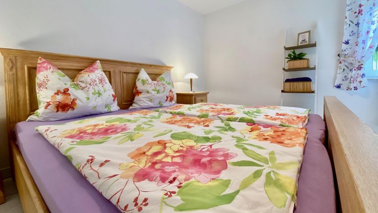 Ein gemütliches Schlafzimmer mit einem Holzbett und bunten Kissen. Die Bettdecke zeigt ein florales Muster und ein Nachtisch steht neben dem Bett.