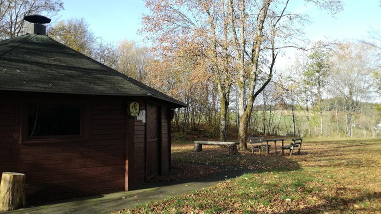 Een klein houten huis omgeven door herfstbomen en kleurrijke bladeren. Op de voorgrond staat een bank langs een pad.