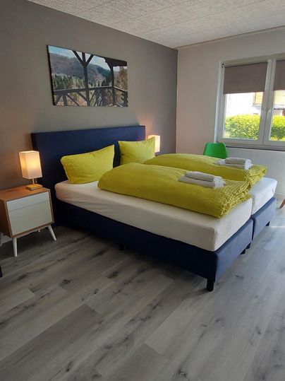 Ein modernes Schlafzimmer mit einem großen, bequemen Bett und frischen, grünen Bettwaren. Es gibt eine Nachtkonsole mit einer Lampe und viel Tageslicht durch das Fenster.