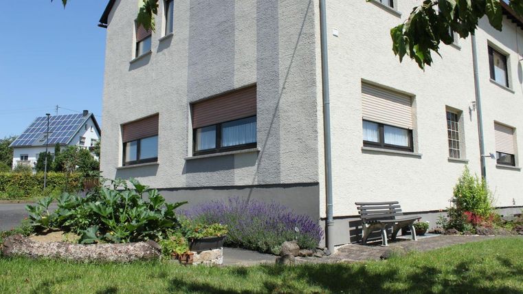 Ein helles Wohnhaus mit einem gepflegten Garten. Im Vordergrund sind Blumen und ein Holzbank zu sehen.