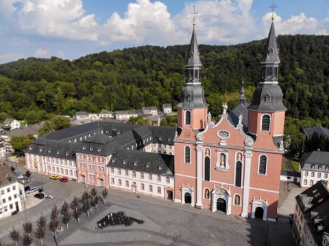 Eine beeindruckende Kirche mit zwei hohen Türmen, umgeben von historischen Gebäuden. Im Hintergrund sind sanfte Hügel zu sehen.