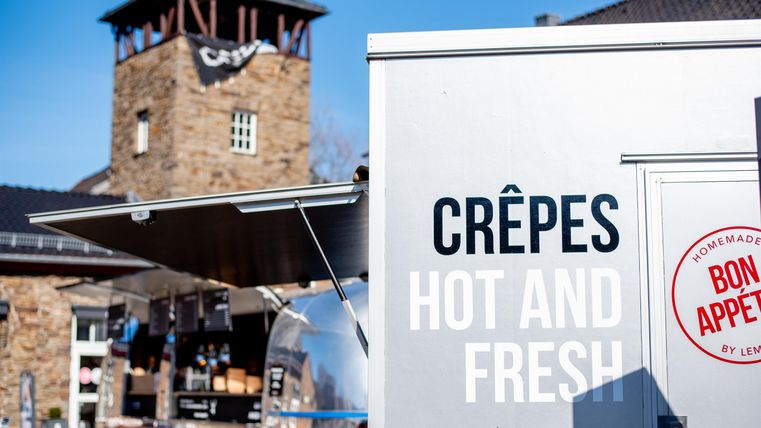Een crêpe-wagen met het opschrift "Hot and Fresh" staat voor een historisch gebouw. De scène geeft een gezellige straateten-sfeer weer.