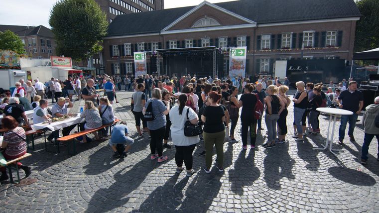 Een grote menigte verzamelt zich op een plein voor een gebouw. Op de achtergrond staan podia en tafels, terwijl de zon schijnt.