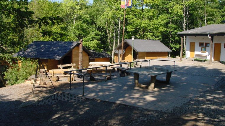 Houten huisjes en een tafeltennistafel buiten, omringd door bomen. Een vlaggenmast met vlaggen staat in het midden.