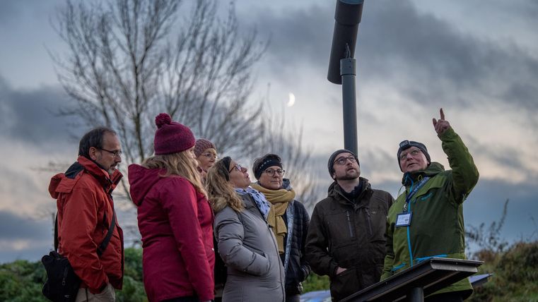 A group of people is standing outside and listening to a speaker who is pointing at a specific point. The sky is cloudy and it is dusk.