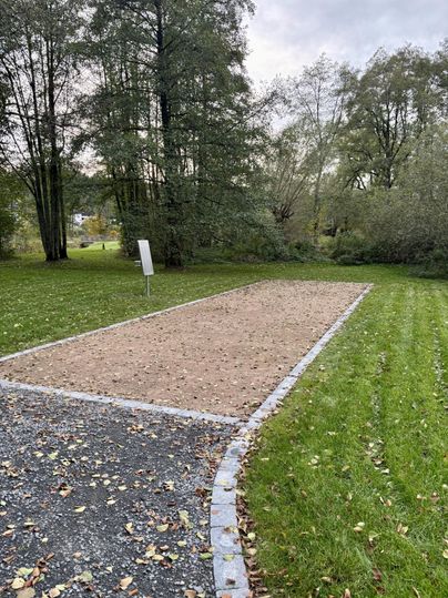 An empty space with sand, surrounded by green areas and trees. In the background, there is a sign.