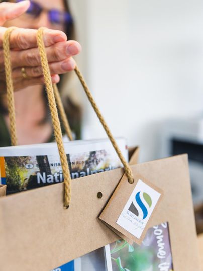 Eine Hand hält eine Papiertüte mit einem Etikett und mehreren Broschüren. Die Tüte hat ein einfaches, elegantes Design.