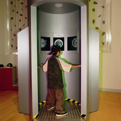 Ein Junge steht in einer interaktiven Ausstellung in einer futuristischen Kabine. Die Wände sind grau, und auf dem Boden sind gelbe Warnlinien sichtbar.