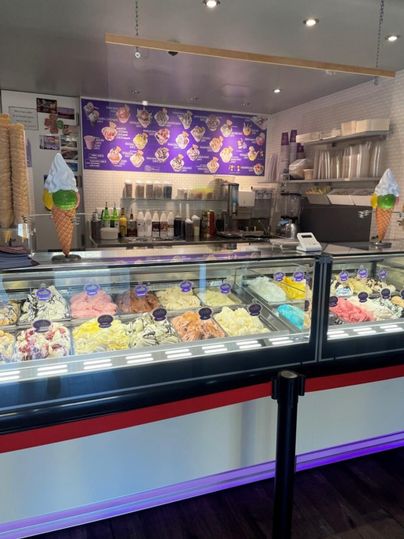 An ice cream parlor with a large variety of colorful ice cream in display cases. In the background, there are tables and chairs, as well as a decorative wall with pictures of ice cream.