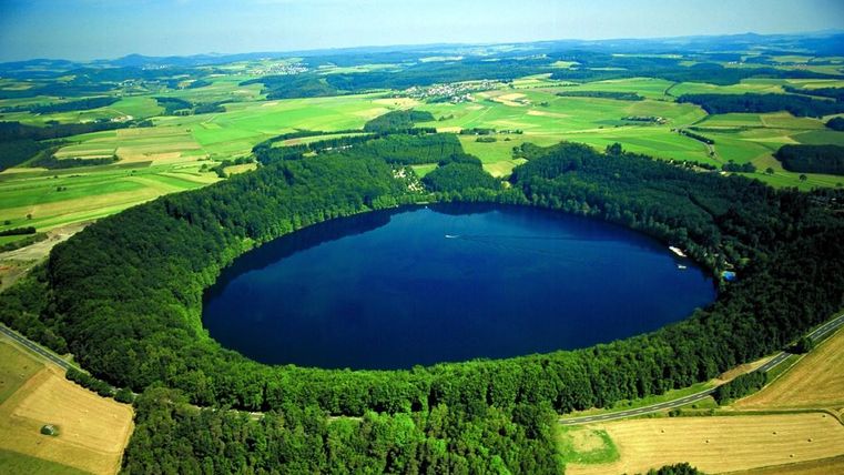 Ein schöner, runder See umgeben von dichten Wäldern und sanften Hügeln. Die klare blaue Wasseroberfläche reflektiert den Himmel.