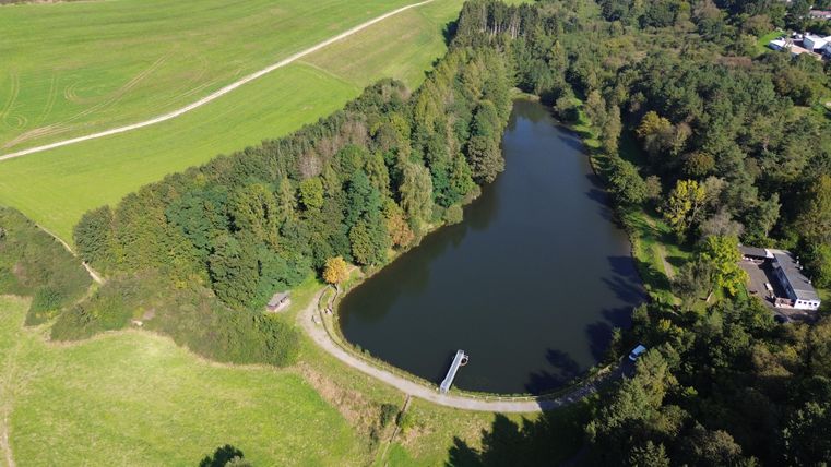 Luchtfoto van het stuwmeer van Gerolstein, omringd door dichte bossen en groene weilanden. Een pad loopt langs de oever, een gebouw is zichtbaar.