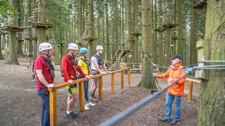 Eine Gruppe von vier Personen in Kletterausrüstung steht in einem Wald. Sie hören einer Sicherheitsanweisung zu, während ein Trainer ihnen zeigt, wie sie einen Seilparcours bewältigen.
