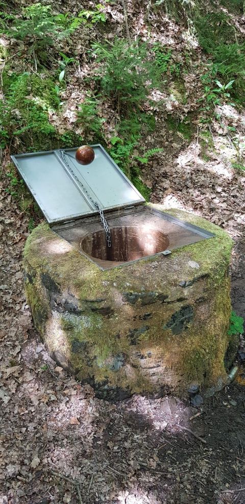 Ein runder Brunnen aus Stein im Wald, mit einem geöffneten Deckel. Wasser fließt aus dem Brunnen und ein Ball schwimmt obenauf.