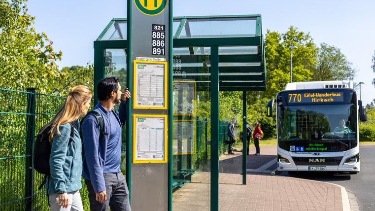 A bus stop with schedules and a passing bus line. Two people are waiting underneath and looking at the displays.