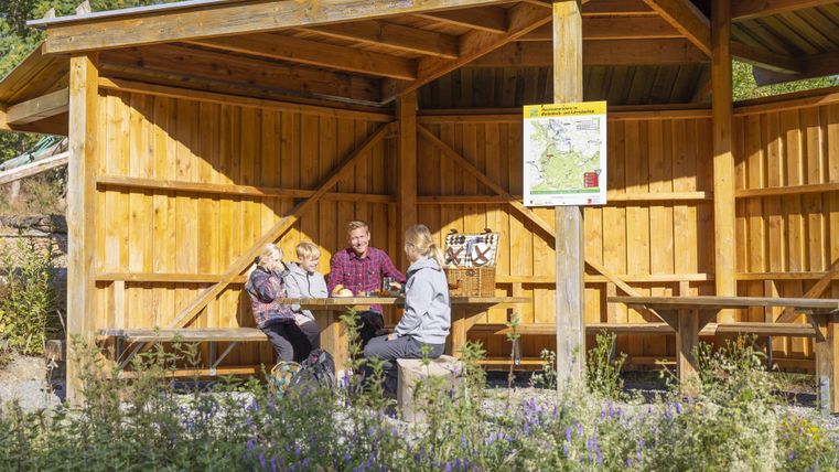 Eine gemütliche Holzunterkunft mit einer Sitzgruppe im Freien. Drei Personen sitzen zusammen und genießen die Zeit im Grünen.