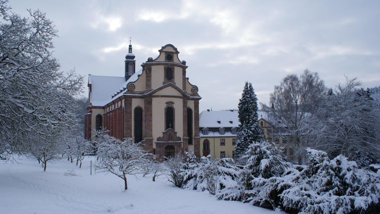 Eine schöne Kirche im Schnee, umgeben von schneebedeckten Bäumen. Der Himmel ist bewölkt, und die Landschaft wirkt friedlich und winterlich.